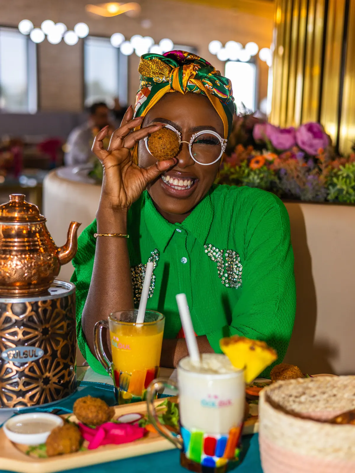 Azeezat at a restaurant table, playfully holding a falafel up to her eye