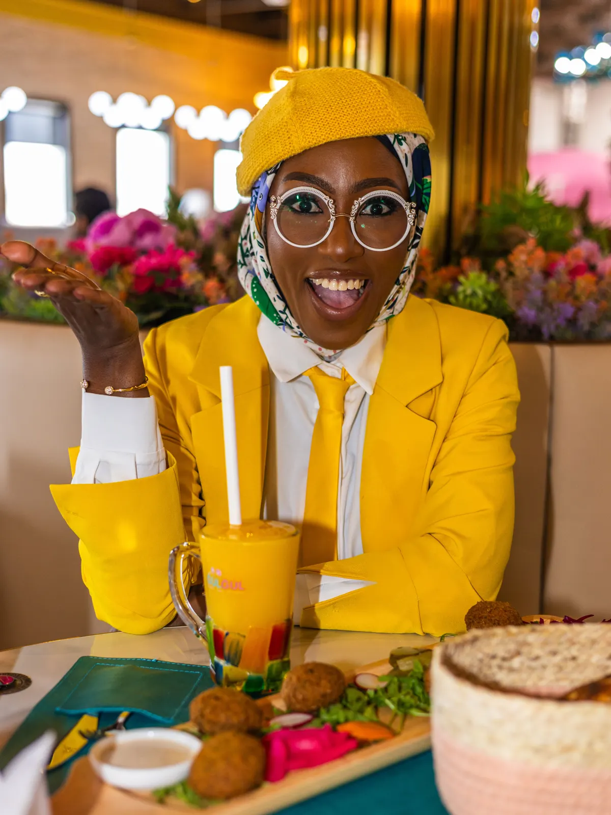 Azeezat in a yellow blazer at a set table with drinks and falafel